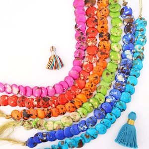 May include: A collection of colorful beaded necklaces in a rainbow of hues, including pink, orange, green, and blue. Each necklace features flat, round beads with a speckled pattern and a gold tassel. A small, multi-colored tassel is also present.