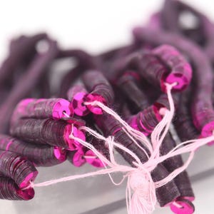 May include: Close-up of a bundle of dark purple sequins with bright pink edges, strung together with pink thread. The sequins are tightly packed, creating a textured appearance. Ideal for crafting and embellishments.