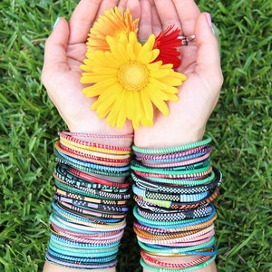 May include: A pair of arms adorned with stacks of colorful bracelets in various patterns and colors, with a backdrop of green grass. The hands hold three gerbera daisies in yellow, orange, and red.