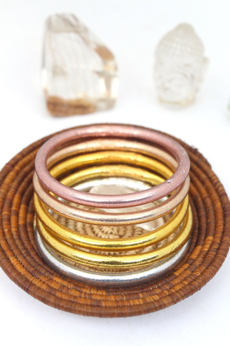 May include: Five stacked metal bangles in a woven basket. The bangles are in shades of rose gold, gold, and silver.