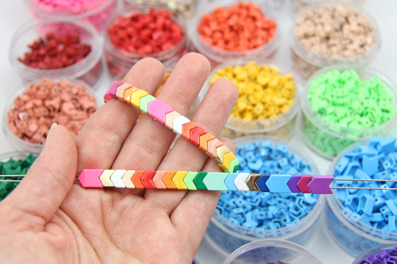 K&ouml;nnte beinhalten: Ein Regenbogenfarbenes Armband aus pfeilf&ouml;rmigen Fliesen mit goldenen Akzenten. Das Armband besteht aus kleinen Keramikfliesen, die auf einer Schnur zusammengef&uuml;gt sind. Die Fliesen sind in einem Regenbogenmuster angeordnet, wobei jede Fliese eine andere Farbe hat. Das Armband wird auf einer Hand gezeigt, wobei die Fliesen zur Kamera zeigen.