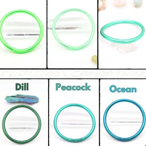 May include: Six colorful bangles in shades of green and blue, including Fluo Green, Spearmint, Green, Dill, Peacock, and Ocean. Each bangle is a solid, circular shape. The names of the colors are written above each bangle.