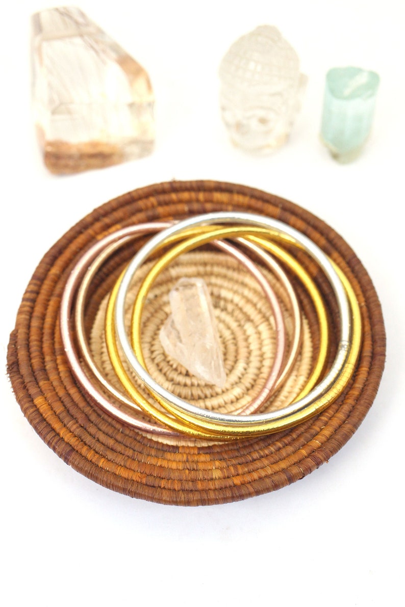 May include: A set of three gold, silver, and rose gold bangles in a woven basket. The basket is brown and has a natural texture.