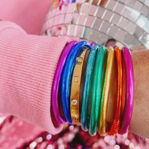 May include: A collection of colorful bangles in a rainbow of hues, including purple, blue, green, gold, orange, and pink. A gold bangle with two small clear stones is also visible. The bangles are stacked on a wrist.