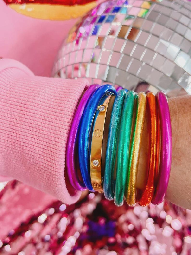 May include: A collection of colorful bangles in a rainbow of hues, including purple, blue, green, gold, orange, and pink. A gold bangle with two small clear stones is also visible. The bangles are stacked on a wrist.