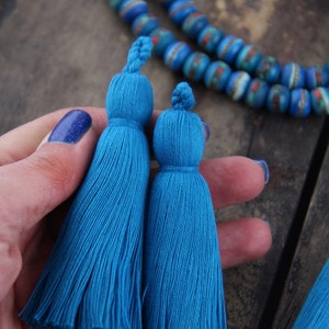 May include: Two blue tassel trim embellishments, one slightly larger than the other, are held in a hand. The tassels are made of soft, fluffy yarn and have a knotted top.