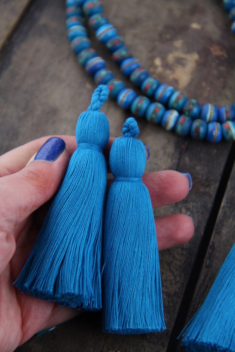 May include: Two blue tassel trim embellishments, one slightly larger than the other, are held in a hand. The tassels are made of soft, fluffy yarn and have a knotted top.