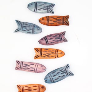 May include: A collection of colorful, carved wooden fish in shades of blue, orange, and purple, arranged on a white background. Also, there are blue and purple beads. These decorative fish are ideal for crafting or jewelry making.