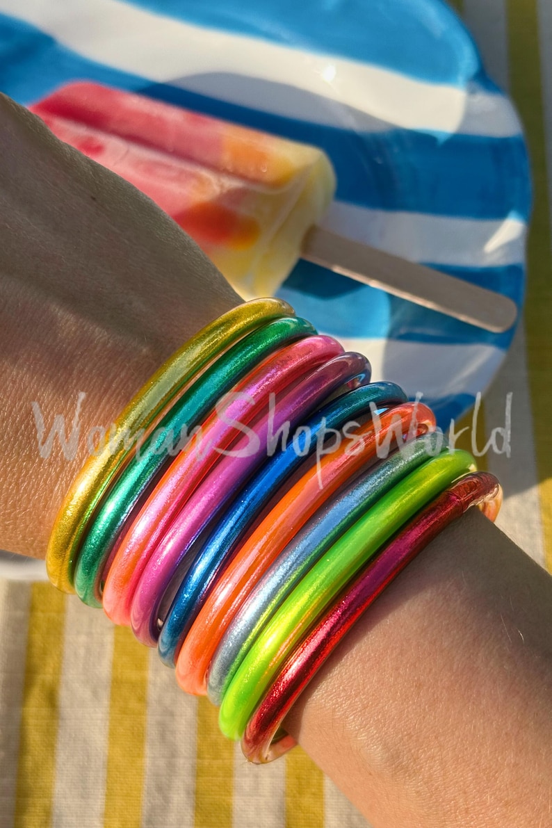 May include: A stack of colorful, metallic bangles in shades of gold, green, pink, blue, orange, silver, and red. The bracelets are stacked on a wrist, with a popsicle and a blue and white striped plate in the background. The text "Woman Shops World" is visible.