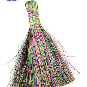 May include: A colorful, multi-colored broom with a green, yellow, and purple handle. The broom is made of natural fibers and has a long, flowing fringe.