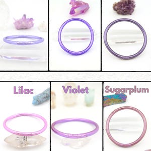 May include: Six colorful bangles in shades of purple, pink, and lavender are displayed. The colors are labeled: Veri Peri, Indigo, Future Dusk, Lilac, Violet, and Sugarplum. Each bangle is a solid color and appears to be made of a flexible material.
