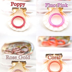 May include: Four seashells each containing a different color of metal rings. The colors are poppy, fluo pink, rose gold, and coral.