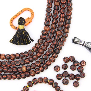 May include: Strands of dark brown wooden beads with red accents and a repeating pattern. The image also shows loose beads, tassels, and an orange carabiner clip. The beads are likely for jewelry making or crafting.
