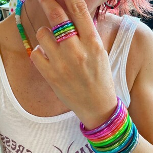 May include: A stack of colorful, iridescent, metallic bangles. The bangles are stacked on a person's wrist and are arranged in a rainbow pattern. The bangles are made of a smooth, shiny material.