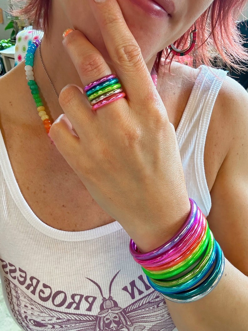 May include: A stack of colorful, iridescent, metallic bangles. The bangles are stacked on a person's wrist and are arranged in a rainbow pattern. The bangles are made of a smooth, shiny material.