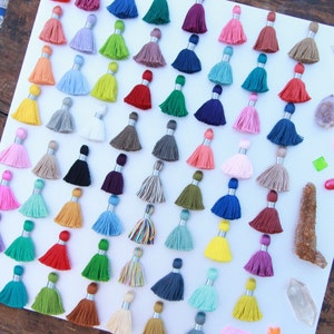 May include: A collection of colorful tassels arranged in rows on a white surface. The tassels are made of various materials, including cotton and silk, and come in a variety of colors, including red, blue, green, yellow, and purple.