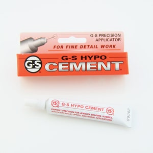 以下が含まれることがあります： 精密なアプリケーターが付いた、赤と白のG-Sハイポセメントのチューブ。チューブには、「G-Sハイポセメント」と「ジュエリー、ビーズ、趣味、時計のクリスタル、光学、産業用途のためのピンポイント精度」というテキストがラベル付けされています。