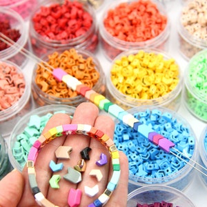 K&ouml;nnte beinhalten: Ein regenbogenfarbenes Armband aus kleinen quadratischen Perlen. Das Armband befindet sich in einer Hand, umgeben von kleinen Plastikbeh&auml;ltern, die mit bunten pfeilf&ouml;rmigen Perlen gef&uuml;llt sind.