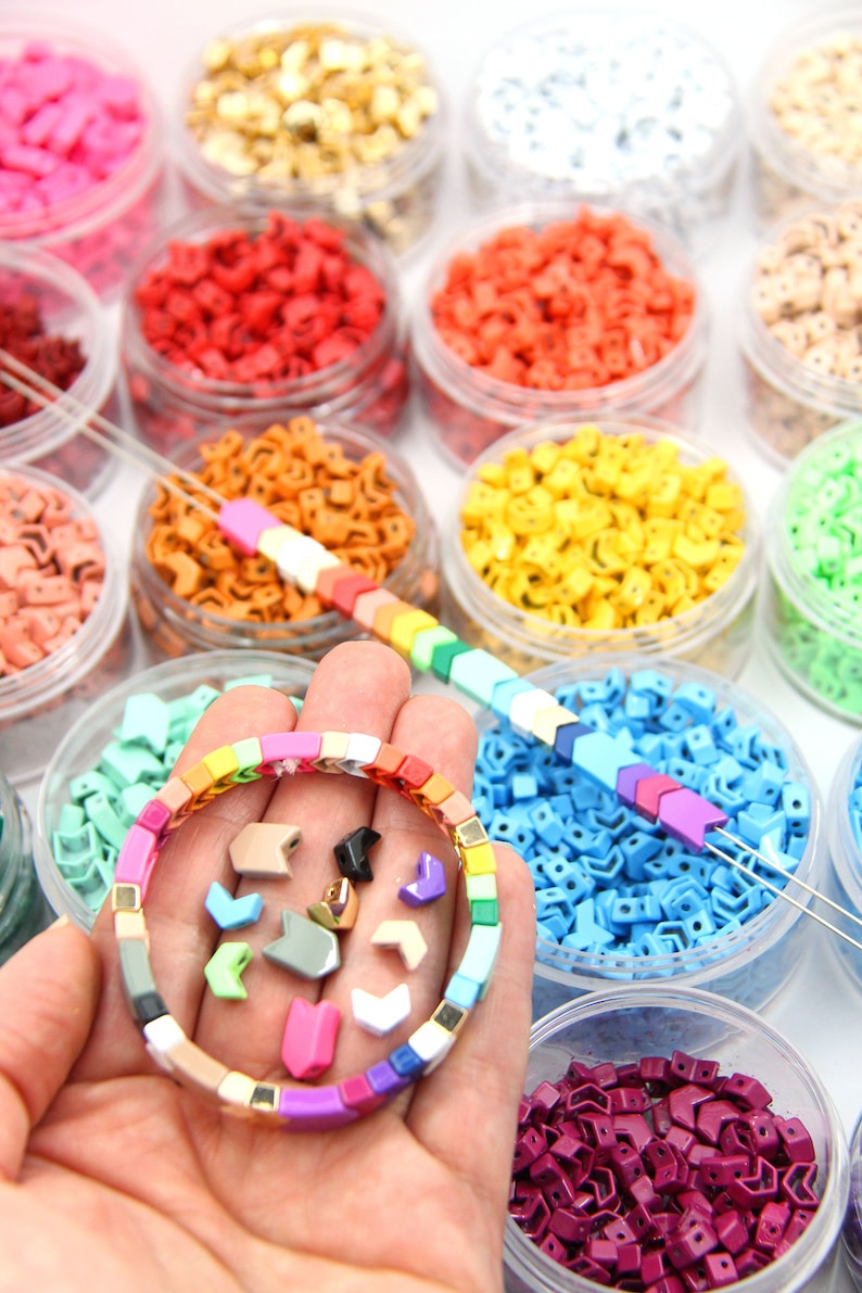 K&ouml;nnte beinhalten: Ein regenbogenfarbenes Armband aus kleinen quadratischen Perlen. Das Armband befindet sich in einer Hand, umgeben von kleinen Plastikbeh&auml;ltern, die mit bunten pfeilf&ouml;rmigen Perlen gef&uuml;llt sind.