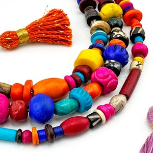 May include: Multicolored wooden bead necklace with a bright orange tassel. The necklace features a variety of shapes and sizes of beads, including round, oval, and cylindrical beads. The beads are made of wood and are painted in a variety of bright colors, including red, orange, yellow, green, blue, and purple.