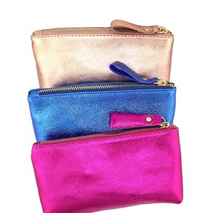 May include: Three metallic zippered pouches in rose gold, blue, and fuchsia. Each pouch has a matching wrist strap and a gold-colored zipper. The pouches are rectangular and appear to be made of a shiny, textured material.