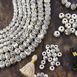 May include: A collection of white and black patterned beads with a gold tassel. The beads are round and have a concentric circle design. The beads are arranged in a pile and a few strands.