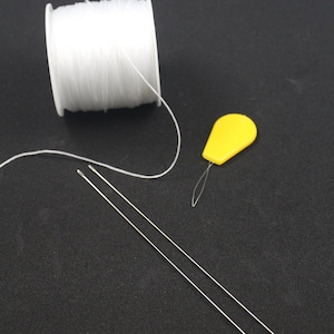 May include: A spool of white thread, two silver needles, and a yellow plastic needle threader on a black surface.