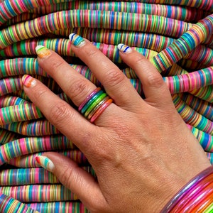 May include: A hand wearing a stack of colorful rainbow rings. The hand is resting on a pile of colorful striped round beads.
