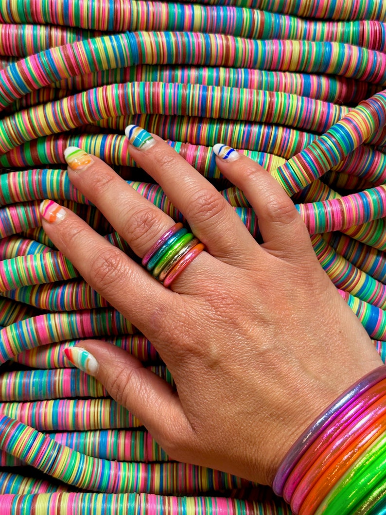 May include: A hand wearing a stack of colorful rainbow rings. The hand is resting on a pile of colorful striped round beads.