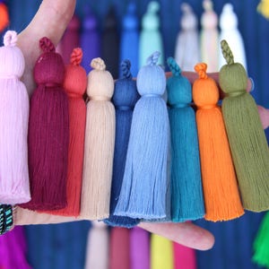 Image shows a hand holding lots of colorful tassels made by WomanShopsWorld. The tassels have a braided loop at the top and nice cotton threads.