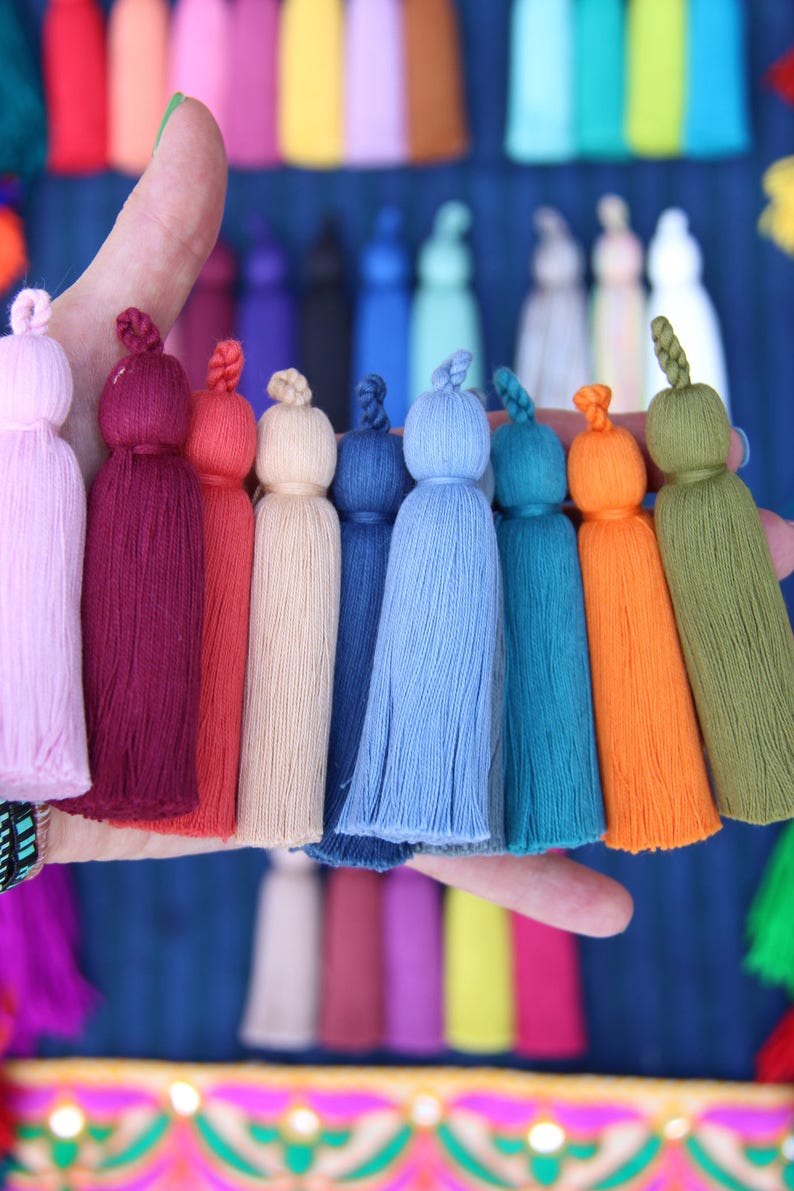 Image shows a hand holding lots of colorful tassels made by WomanShopsWorld. The tassels have a braided loop at the top and nice cotton threads.