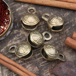 May include: Five small, gold-coloured bells with intricate designs. The bells have a round shape and a small loop at the top for hanging. The bells are arranged on a wooden surface.