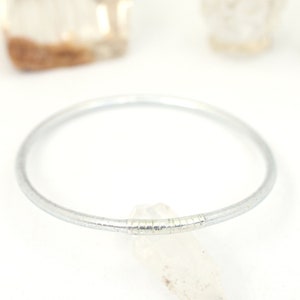 May include: A silver bangle bracelet with a textured design. The bracelet is lying on a white surface with a clear crystal next to it.
