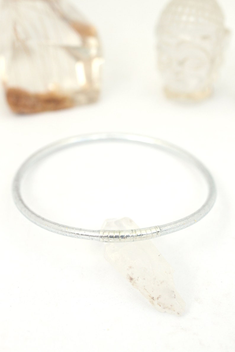 May include: A silver bangle bracelet with a textured design. The bracelet is lying on a white surface with a clear crystal next to it.