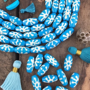 May include: A collection of turquoise blue beads with white floral designs, along with matching tassels and star-shaped accents. The beads are oval-shaped and strung together, suggesting jewelry components or craft supplies.