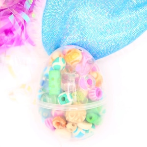 Puede incluir: Un huevo de Pascua de plástico transparente lleno de cuentas de colores. Las cuentas son de varias formas y tamaños, incluyendo redondas, ovaladas y en forma de corazón. El huevo está sobre una superficie blanca.