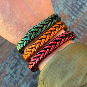 May include: Three braided bracelets in shades of green, orange, and red. The bracelets are worn on a wrist, showcasing their metallic sheen. The braided design is a prominent feature, adding texture and visual interest to the jewelry.