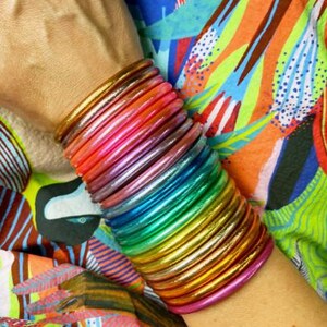 May include: A stack of colorful, rainbow-hued bangles. The bangles transition through shades of pink, orange, yellow, green, and blue. The bracelets are stacked on a wrist, with a vibrant, patterned fabric in the background.