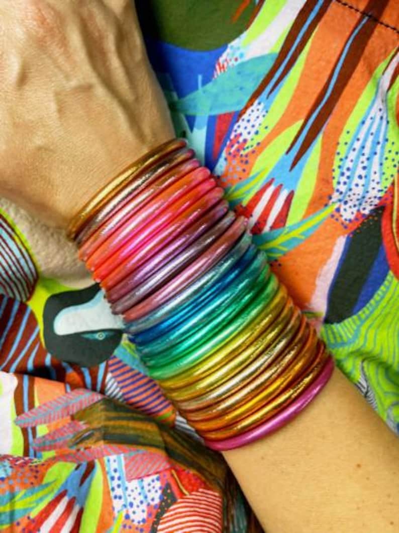 May include: A stack of colorful, rainbow-hued bangles. The bangles transition through shades of pink, orange, yellow, green, and blue. The bracelets are stacked on a wrist, with a vibrant, patterned fabric in the background.