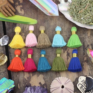 May include: Assortment of 10 colorful cotton tassels with gold trim. The tassels are arranged in two rows of five on a wooden surface.