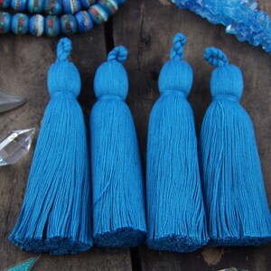 May include: Four teal blue cotton tassels with a knotted top. The tassels are arranged in a row on a wooden surface.