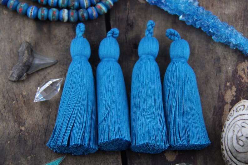 May include: Four teal blue cotton tassels with a knotted top. The tassels are arranged in a row on a wooden surface.