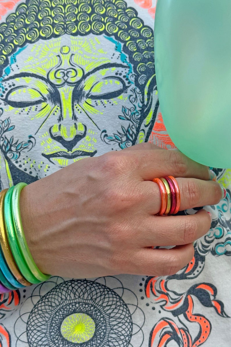 May include: A hand wearing a stack of colorful rings, with a green, yellow, and white patterned shirt in the background.