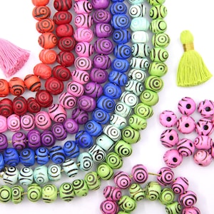 May include: A rainbow assortment of colorful wooden beads with black concentric circles painted on them. The beads are strung together in a long strand and there are smaller strands of beads in pink and green.