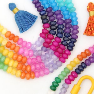 May include: A rainbow of colorful gemstone beads strung together on a necklace. The beads are faceted and have a smooth, polished finish. The necklace is accented with a yellow metal clasp and two tassels, one blue and one orange.
