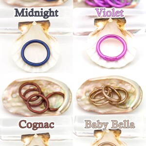 May include: Four sets of colorful metal rings in different colors, including midnight blue, violet, cognac, and baby bella, displayed in seashells. The rings are arranged in a single row, with each set in its own seashell.