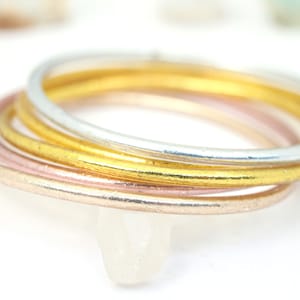 May include: A set of four thin metal rings in gold, silver, and rose gold. The rings are stacked on top of each other.