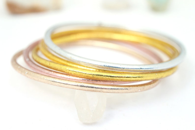 May include: A set of four thin metal rings in gold, silver, and rose gold. The rings are stacked on top of each other.