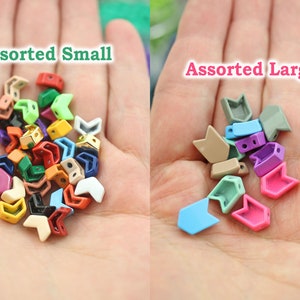 K&ouml;nnte beinhalten: Sortiertes kleine und gro&szlig;e pfeilf&ouml;rmige Metallperlen in verschiedenen Farben. Die Perlen sind in zwei Haufen auf der Hand einer Person angeordnet. Der Text "Assorted Small" befindet sich &uuml;ber den kleineren Perlen und der Text "Assorted Large" &uuml;ber den gr&ouml;&szlig;eren Perlen.