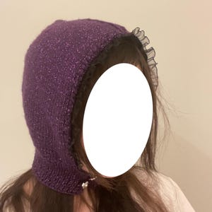 May include: A purple knitted hood with a sparkly finish and a black lace trim. The hood features a small decorative jewel near the face opening. The hood is designed to be worn on the head.